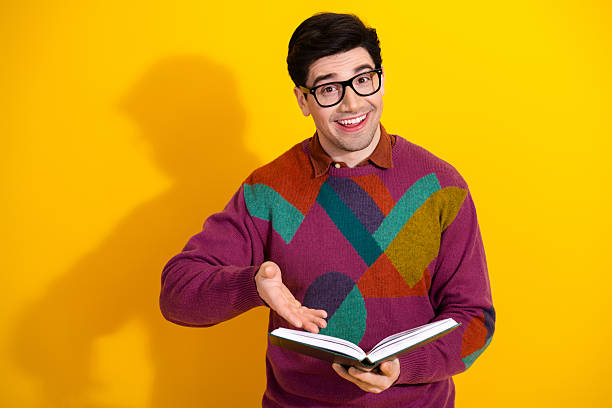istock Young man wearing stylish sweater and glasses holding an open book while smiling against a vibrant yellow background 2207140063