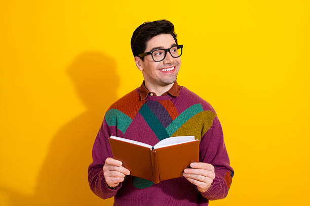 istock Stylish young man with glasses smiling while holding a book against a vibrant yellow background 2207140102