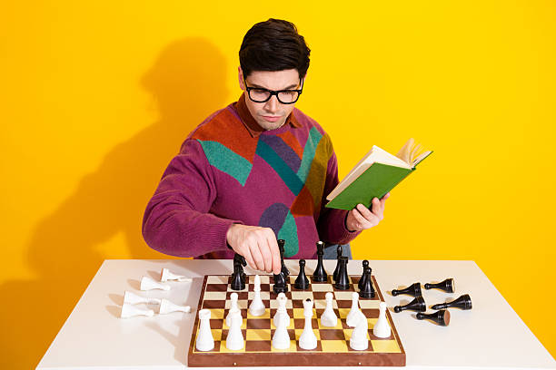 istock Young man reading a book and analyzing chess pieces at a table against a bright yellow background 2207140229