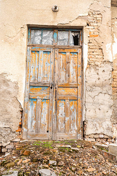 This image features an old, weathered wooden door with peeling paint in shades of blue and orange. The door is framed by crumbling walls and exposed brick, adding a sense of abandonment and decay. The cracked glass window and rugged surroundings emphasize the aged character of the structure, making it perfect for projects that evoke rustic, vintage, or dilapidated aesthetics.