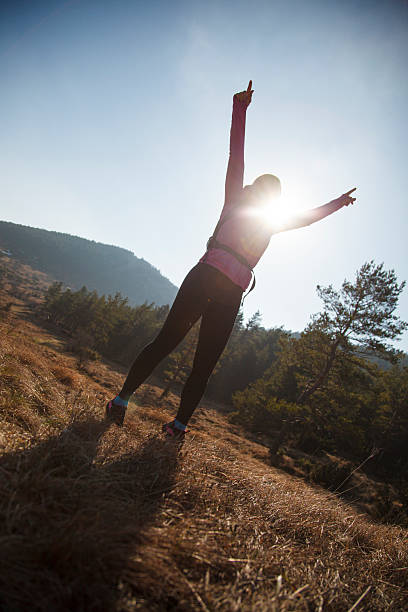 A female trail runner in sportswear raises her arms in victory, standing on a grassy hillside with the sun shining behind her. The golden sunlight creates a dramatic silhouette effect. The outdoor setting features rolling hills, pine trees, and a clear blue sky, capturing the essence of freedom, achievement, and personal triumph.