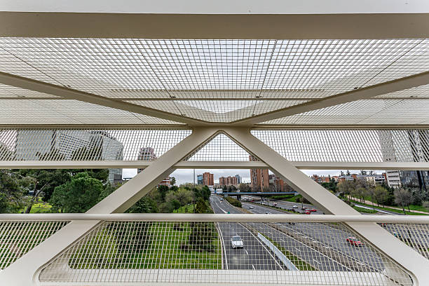 Madrid. Spain. Offering a captivating view from a modern white metal bridge with a grid design, overlooking a bustling highway and serene city park