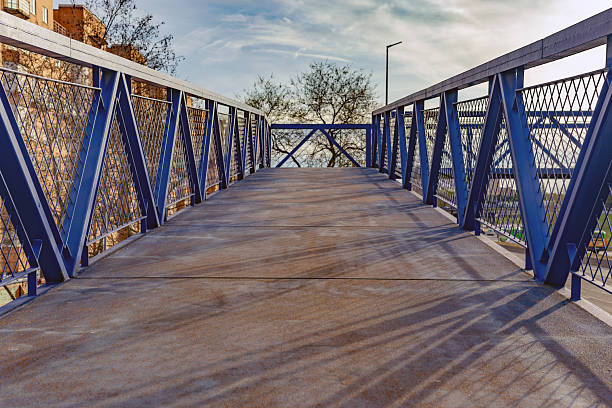 Madrid. Spain. Low angle view of a modern pedestrian bridge featuring a blue steel truss frame and a concrete floor, leading towards a cloudy sky