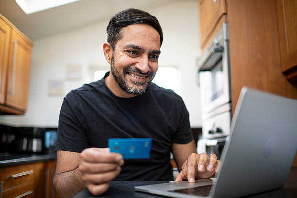 istock Cheerful man holding a credit card and making an online payment on a laptop in the kitchen at home 2207149708