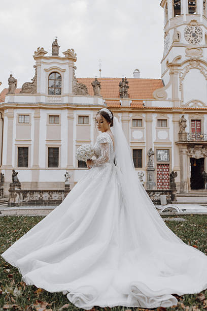 A bride in a long white gown stands in front of a building with a clock tower. She is holding a bouquet of flowers