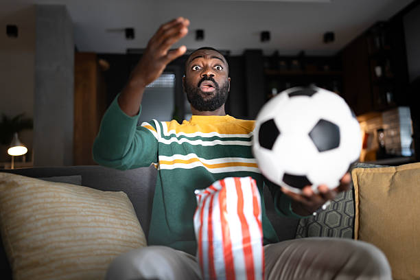 istock A surprised man reacts dramatically while watching a soccer match on TV at home. He sits on a couch with a bowl of popcorn as a soccer ball seemingly comes toward him, adding excitement to the moment. 2207151178