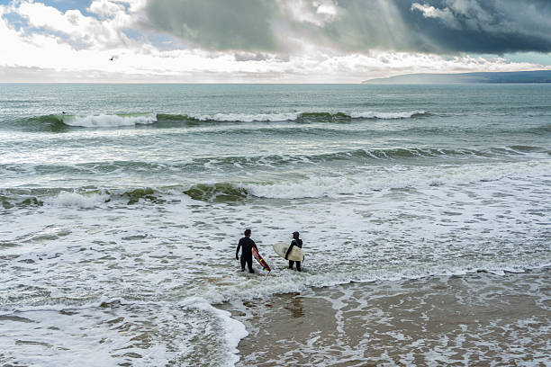 West Cliff Beach, Bournemouth, UK - February 24th 2024: Two surfers holding their surfboards entering the sea.