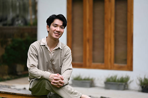 An Asian man sits on a wooden bench in a comfortable pose, smiling gently, contentment, and an easygoing lifestyle, perfect for promotions related to well-being or outdoor retreats.