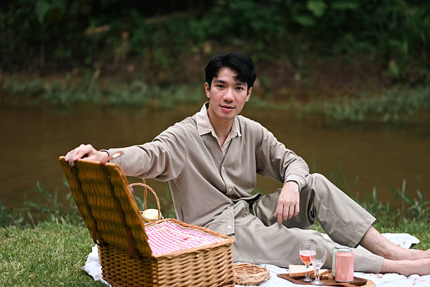 A man sits by the riverside, preparing for a peaceful picnic. He opens a wicker basket filled with food and drinks, enjoying a tranquil outdoor moment.