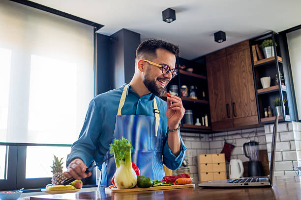 istock A young, smiling man in the kitchen is cutting vegetables on a board while attentively following a recipe on his laptop 2207167840