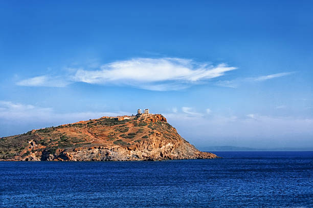 The Temple of Poseidon on a rock rises above the blue sea. A white cloud in a blue sky, like a hat on a temple. Beautiful seascape. Europe, Greece, Cape Sounion, Aegean Sea