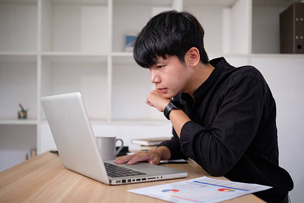 istock Young businessman in a black shirt intently works on his laptop, analyzing data and documents on his desk. 2207192953