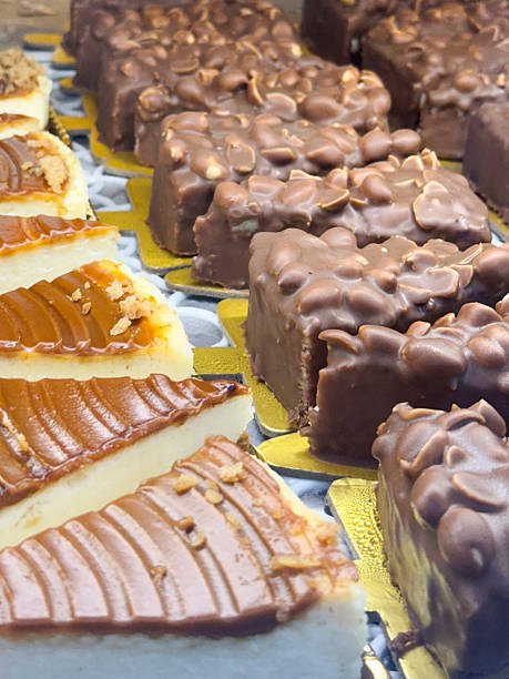istock Full frame image of bakery display of indulgent desserts in rows behind glass panel, shelf containing toffee caramel cheesecakes  and chocolate peanut slices on individual golden, triangular trays, elevated view, focus on foreground 2207199760