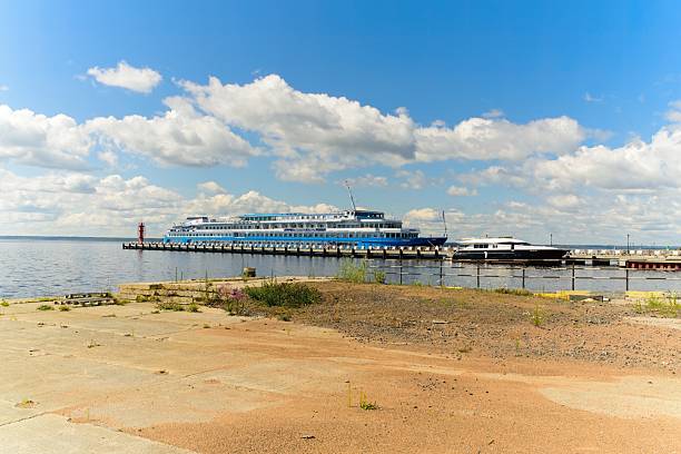Konevets Island, Russia, July 13, 2024.Ships docked at a pier on a lake under a clear blue sky with fluffy clouds. A large blue and white vessel and a smaller boat are moored alongside the pier, which extends out into the water, marked by a red lighthouse. The foreground features a sandy and grassy shore area, while the background shows the expanse of the lake meeting the horizon. The setting is likely serene with a summery atmosphere.