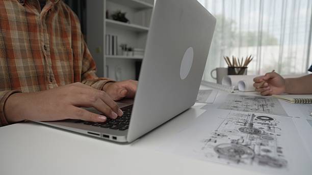 istock Person Working on Laptop with Technical Drawings on Table in Office Space 2207210579