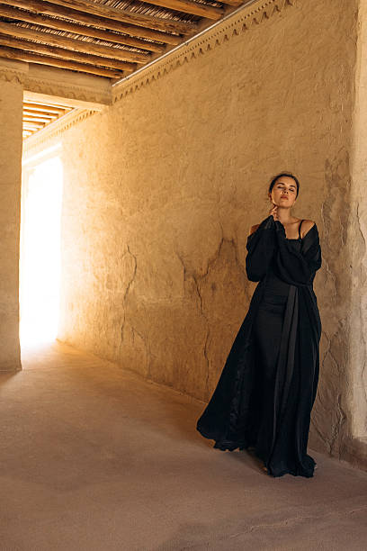 The image captures a serene woman dressed in black attire in an architectural hallway. The setting features textured walls, warm lighting, and detailed ceilings, highlighting the rich cultural and artistic ambiance.