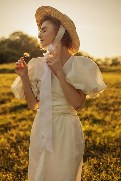 This evocative image depicts a young woman with curly red hair walking through a sunlit meadow during late afternoon. Her vintage-style white dress, with puffed sleeves and a tie at the neck, adds a timeless elegance to the scene. She appears absorbed in the moment, gently touching the wildflowers as she walks by. The warm, natural lighting and the open field background enhance the pastoral and peaceful ambiance, evoking a sense of freedom and connection to nature.