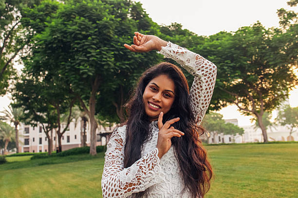 A young woman poses gracefully in a green park, her arms elegantly framing her face as she smiles warmly. Dressed in a delicate white lace outfit, she radiates charm and confidence against the serene backdrop of lush trees and open space. The soft natural light enhances her glowing complexion, adding a touch of magic to this lighthearted and artistic moment.
