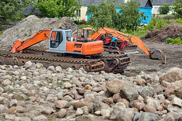 A large excavator with tracks sits partially submerged in a river. The machine is parked on a bed of large rocks near the shore.