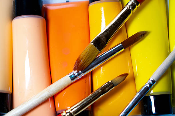 Tubes of acrylic paint in various shades of red and pink are arranged side by side. Two paintbrushes with red handles and golden ferrules cross over the tubes. The brush bristles appear clean and dry, suggesting they are ready for use. The smooth, shiny surface of the paint tubes contrasts with the soft texture of the brushes, creating a visually appealing composition ideal for artistic inspiration.