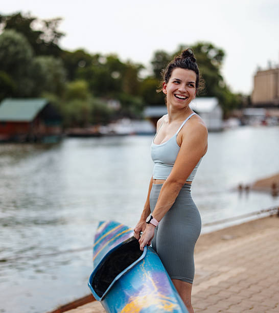 A cheerful woman stands by a river, holding a kayak, radiating joy and energy, portraying fitness and outdoor enthusiasm with a serene water body and lush trees in the background.