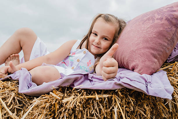 A cute little girl is lying on a straw bed covered with a purple bedspread. The girl is wearing a white dress. She is very happy to spend time in nature. The girl holds her finger up