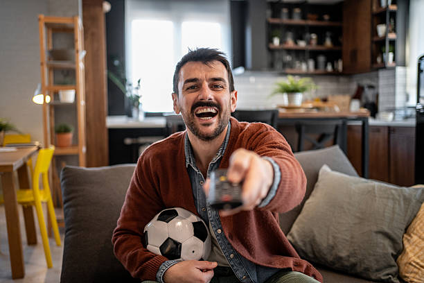 istock Excited soccer fan watching tv and holding remote control 2207250353
