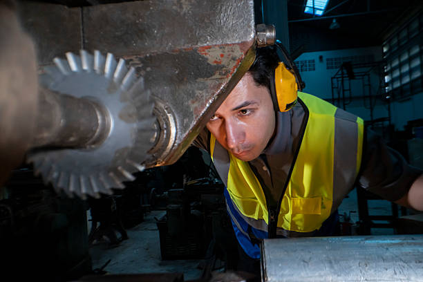 industrial engineer intently operates a gear driven machine in a workshop. Wearing ear protection and a reflective vest, he focuses on precision work, ensuring safe operation of the heavy equipment.