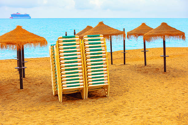 Beach scene with neatly stacked lounge chairs surrounded by straw umbrellas, ready to welcome visitors. Calm sea stretches out to horizon, large ship peacefully sails under a soft sky