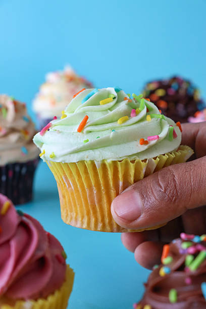 istock Close-up image of individual vanilla cupcake with mint green icing held by unrecognisable person, homemade, cupcakes in paper cake cases, decorated with butter icing piped swirls and hundreds and thousands, turquoise blue background, focus on foreground 2207288987