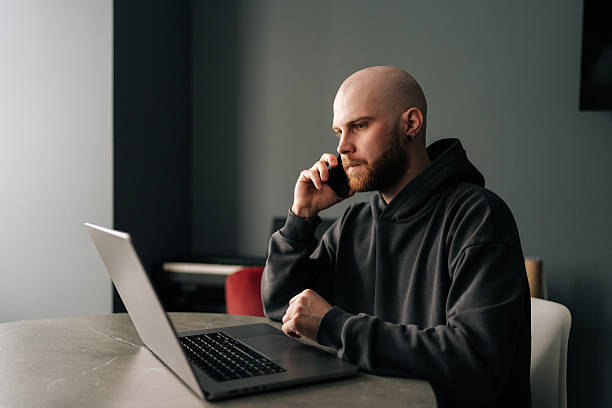 istock Focused young bald freelancer with beard wearing dark hoodie sitting at table, talking on mobile phone and working on laptop computer in modern apartment 2207294888