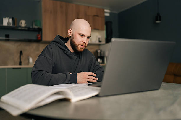 istock Focused bald young man in hoodie taking notes sitting at kitchen table with laptop computer, managing online business and enjoying comfort of working from home. 2207294890