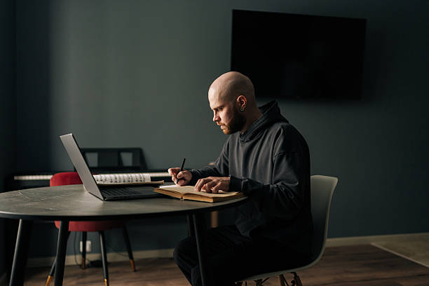 istock Side view of focused young man in hoodie taking notes sitting at table with laptop computer, managing online business and enjoying comfort of working from home 2207294901