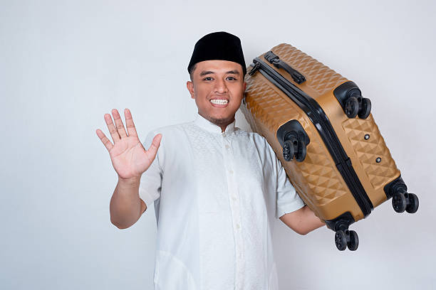 istock Smiling Asian Muslim man wearing white prayer clothes standing on isolated white background while carrying suitcase for mudik (going back to hometown) 2207297621