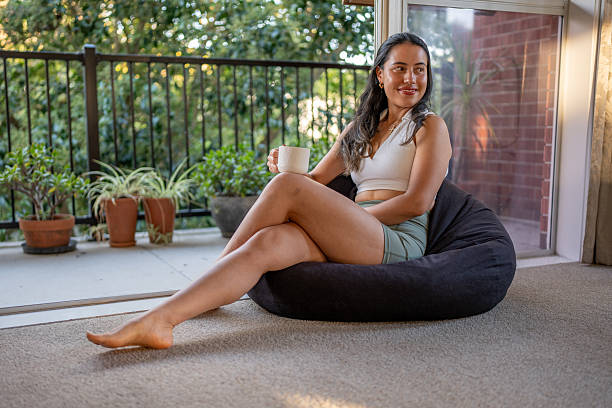 A young woman sits on a bean bag near a balcony door, holding a white mug