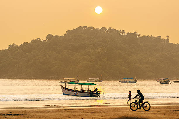 An atmospheric beach scene depicting a tranquil sunset in Goa, India, featuring boats on calm waters and people enjoying a bicycle ride along the shore.