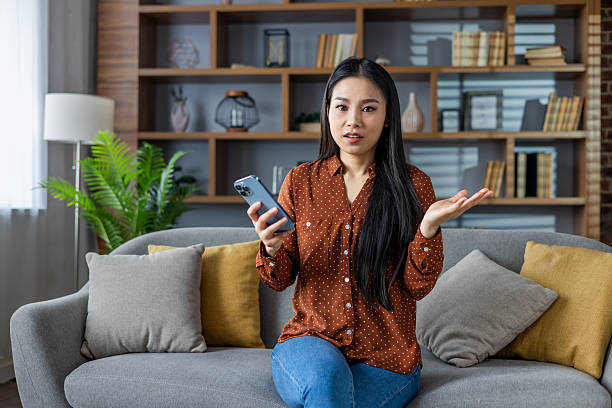 Asian woman sits on sofa holding smartphone with questioning expression. Her casual attire and home setting create relaxed atmosphere. Bookshelves and pillows add comfort to living room scene.