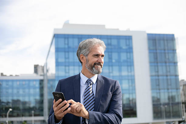 istock Middle-Aged Bearded Businessman with Smartphone in Front of Office Building 2207315740