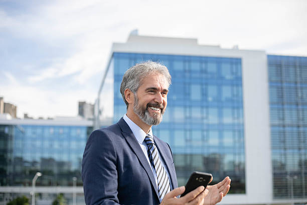 istock Middle-Aged Bearded Businessman with Smartphone in Front of Office Building 2207315768
