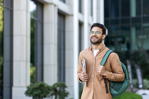 istock Student walking outside university campus with books in hand. Young man with backpack in casual clothes, happy and smiling 2207319194