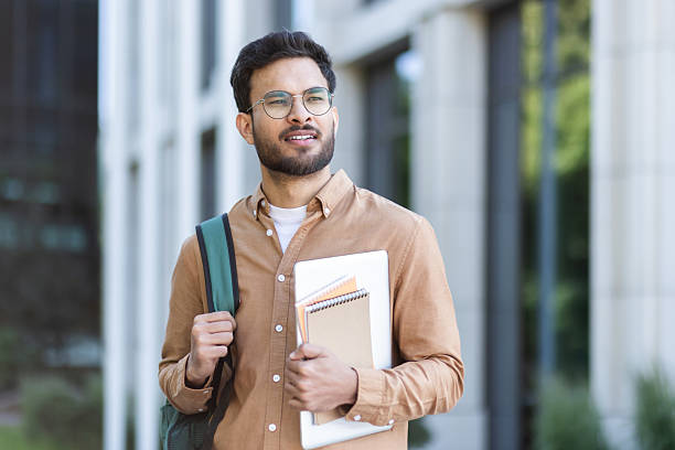 istock Student walking outside university campus with books in hand. Young man with backpack in casual clothes, happy and smiling 2207319197