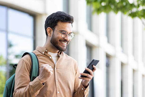 Man outside university campus with phone in hand celebrating successful exam results, student with backpack holding hand up in triumph gesture, received online notification on smartphone