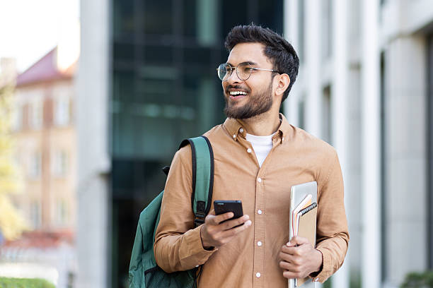 istock A happy student in glasses is walking outdoors and holding a laptop, notebooks, and phone while wearing a backpack. 2207319205