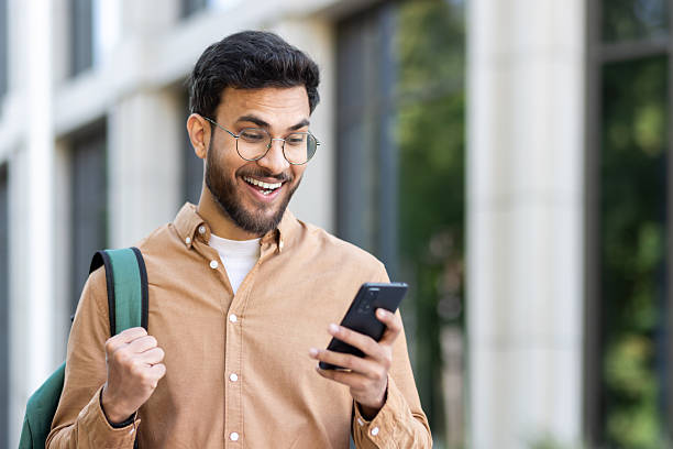 istock Man outside university campus with phone in hand celebrating successful exam results, student with backpack holding hand up in triumph gesture, received online notification on smartphone 2207319210