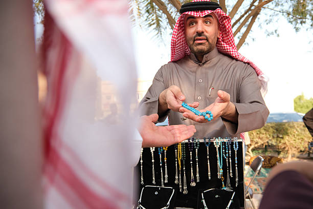 A mid adult trader in traditional Saudi clothing is selling prayer beads and other jewelry at the market. He is showing some blue prayer beads to potential customers