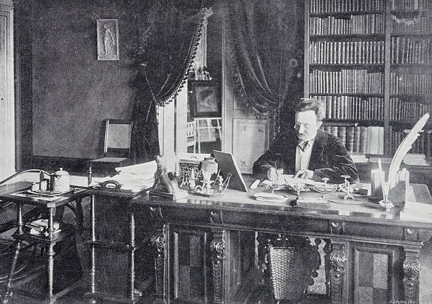 istock Man sitting at his desk in his library writing a letter, in the background full bookshelves and an easel with a framed female portrait 2207321681