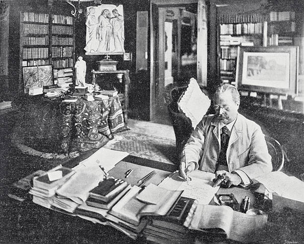 istock Man sitting at his desk in his library writing a letter, in the background full bookshelves and a round table with a white statue and maps 2207321698