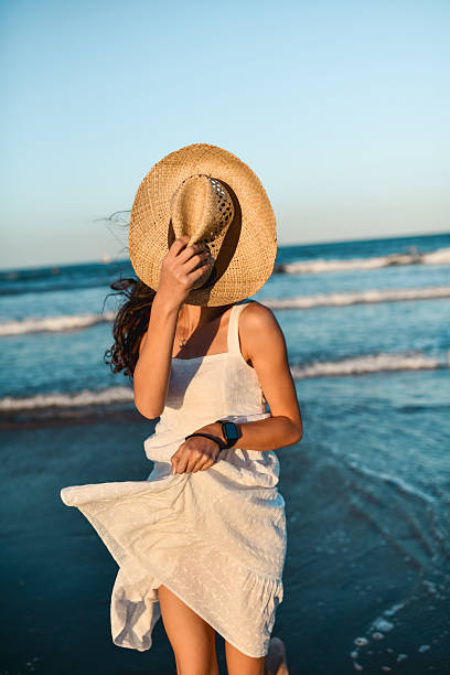 Female Hiding Face With Sun Hat While Walking Next to Ocean in Flowy White Dress