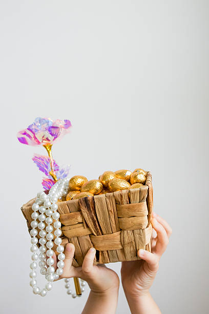 Child holding wicker basket with gold eggs, beads, and pink flower.