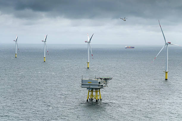 A wide shot shows an offshore wind farm on a choppy, grey sea under a dense, overcast sky. Multiple white wind turbines with red markings on their blades are spread across the water, each mounted on a yellow foundation. In the foreground, a grey and yellow platform structure is visible. A helicopter flies in the sky above the wind farm, and a large cargo ship is seen in the distance on the horizon. The scene illustrates a substantial wind energy generation facility in a marine environment.